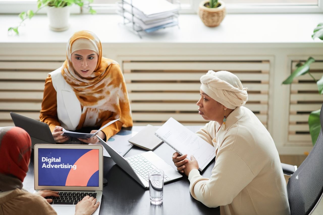 Services Three women in hijabs collaborating on digital advertising strategies around a table with laptops.